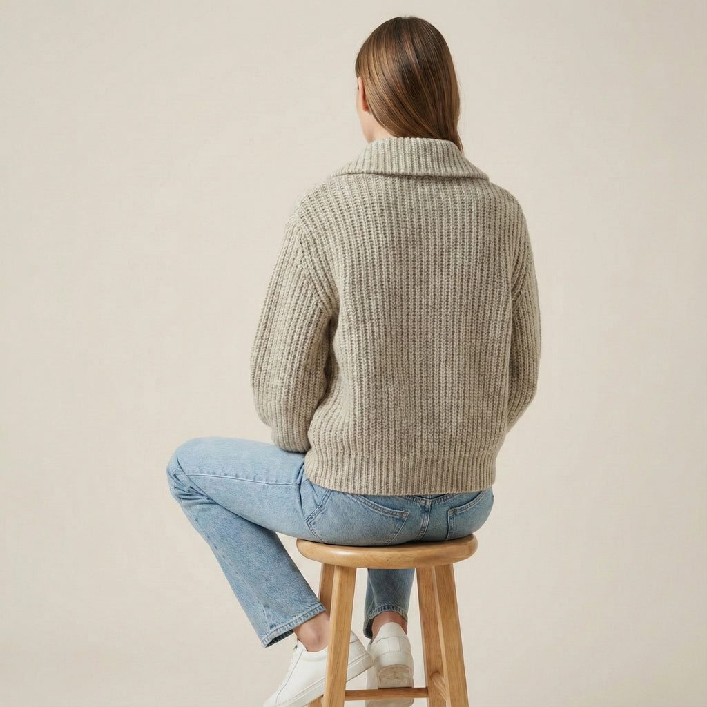 Person wearing a beige knitted sweater and blue jeans sitting on a wooden stool against a plain background