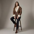 Woman wearing a brown leather coat with white fur trim, sitting on a wooden stool against a plain background.