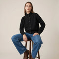 Woman wearing a black sweater and blue jeans sitting on a wooden stool against a beige background