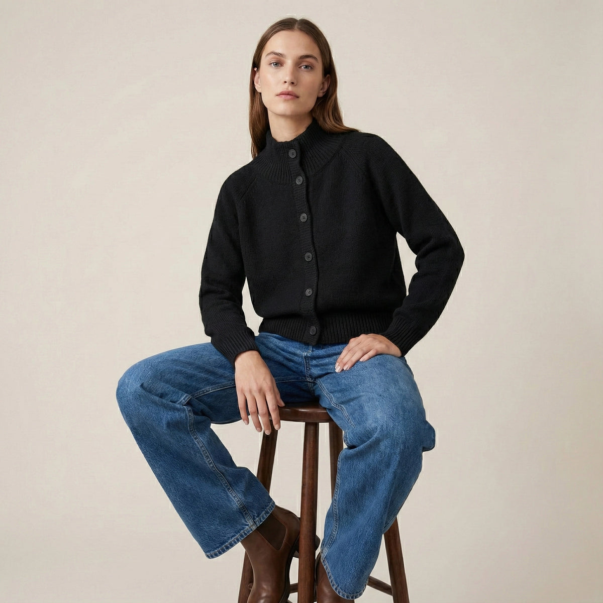 Woman wearing a black sweater and blue jeans sitting on a wooden stool against a beige background