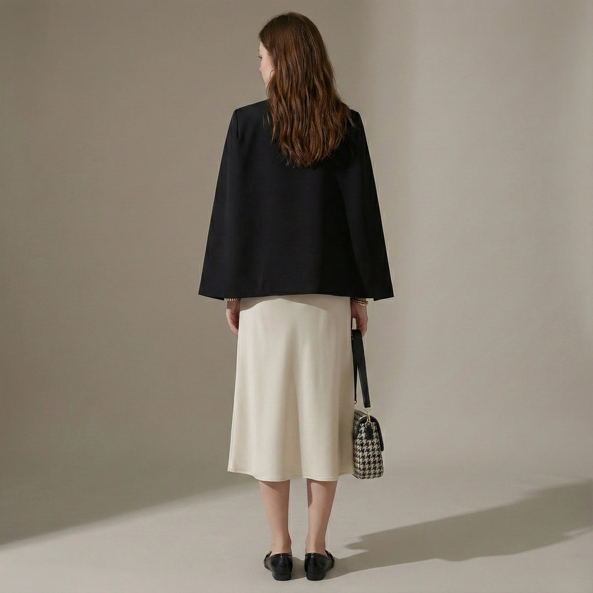 Woman wearing a black coat and beige skirt holding a checkered bag against a plain background