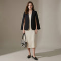Woman in a black blazer and beige dress holding a checkered handbag against a neutral background