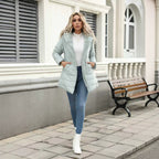 A woman stands on a paved sidewalk in front of a building, wearing a light sage green puffer jacket over a white top and blue jeans, with white ankle boots. A wooden bench is visible in the background.