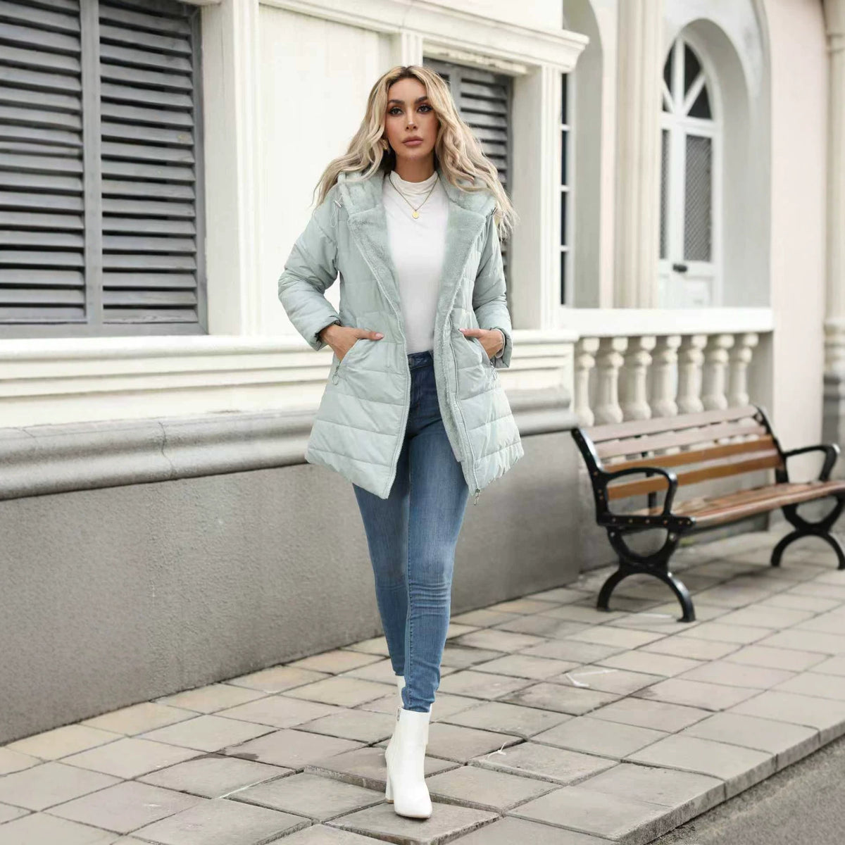 A woman stands on a paved sidewalk in front of a building, wearing a light sage green puffer jacket over a white top and blue jeans, with white ankle boots. A wooden bench is visible in the background.
