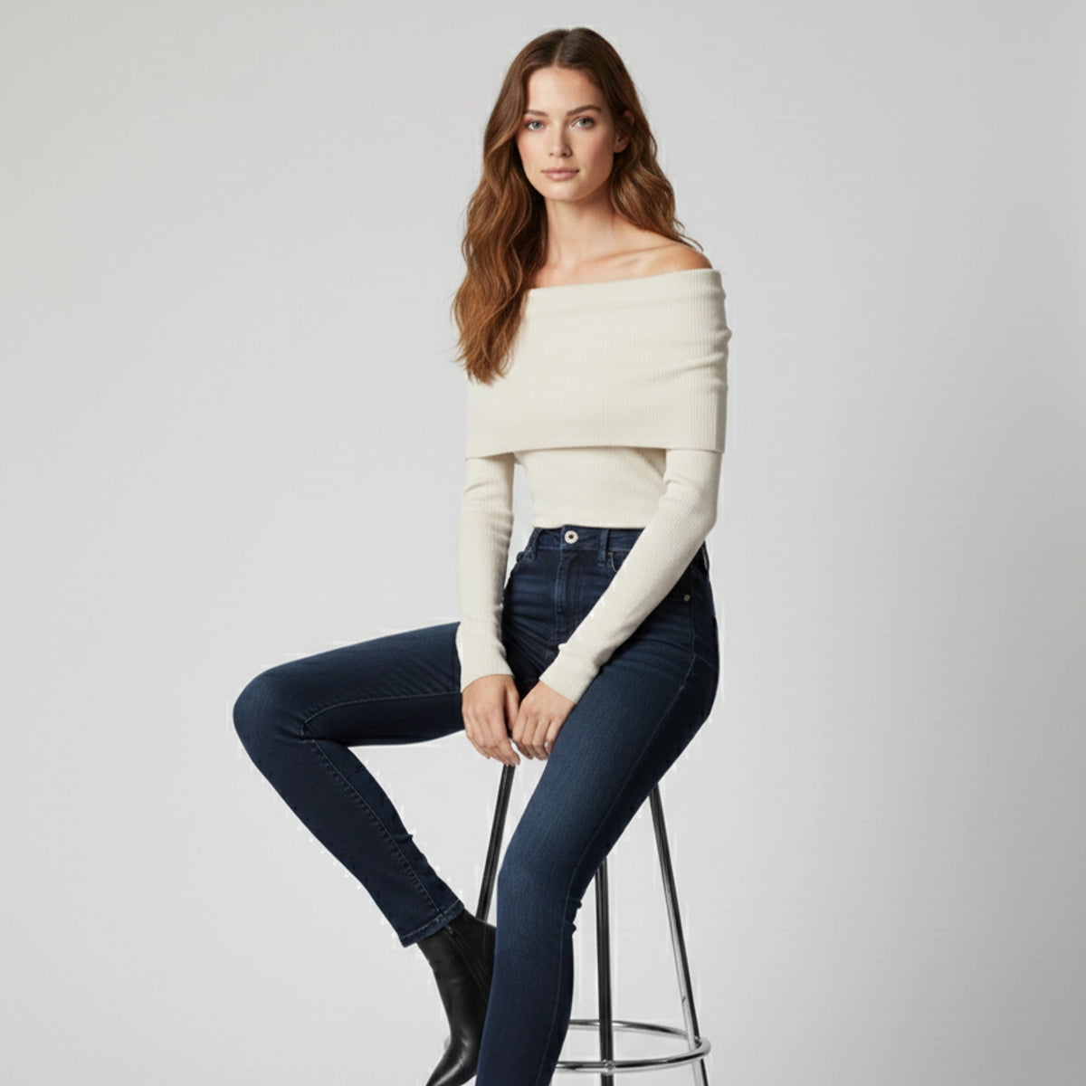 Woman wearing a white off-shoulder sweater and dark jeans sitting on a stool against a plain background