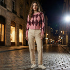 Woman wearing a pink argyle sweater and beige pants standing on a city street at night.