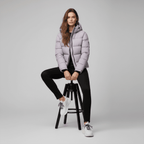 Woman wearing a light purple puffer jacket sitting on a black stool against a gray background
