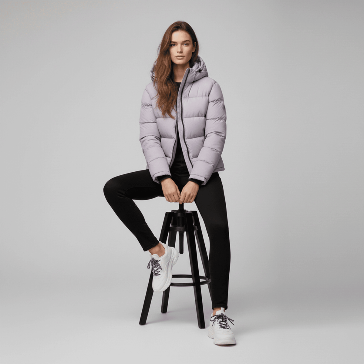 Woman wearing a light purple puffer jacket sitting on a black stool against a gray background