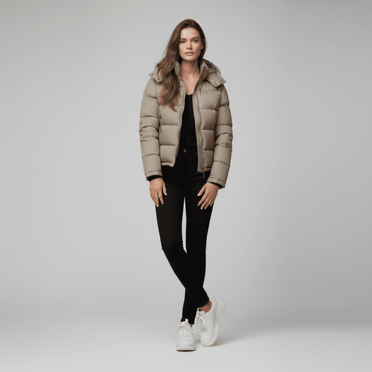 Woman wearing a beige puffer jacket, black pants, and white sneakers on a gray background