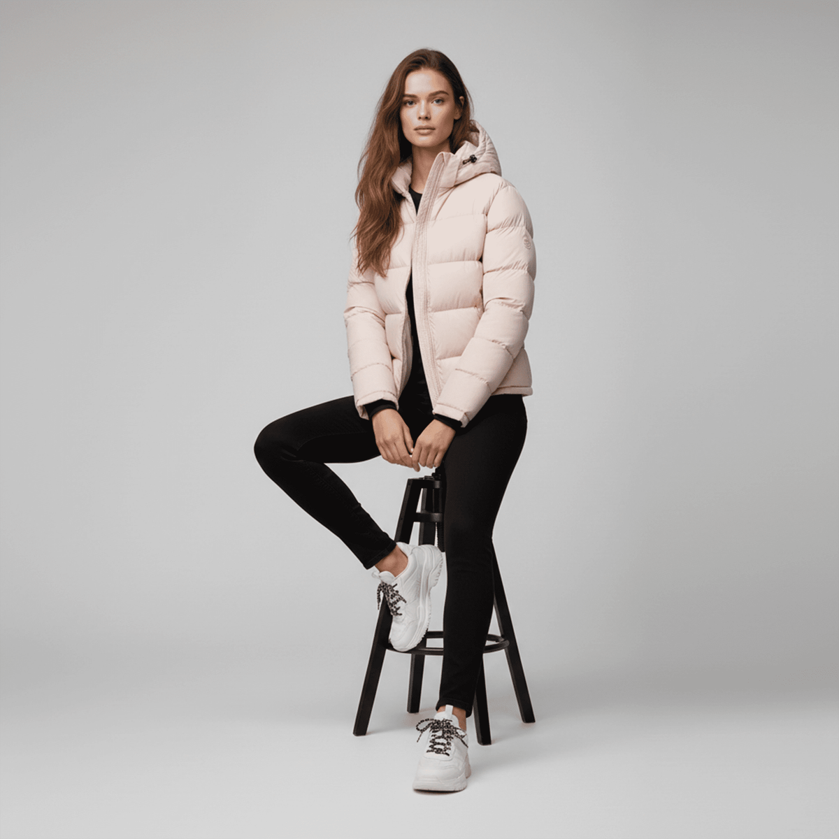 Woman wearing a pink puffer jacket sitting on a black stool against a gray background