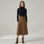Woman wearing a black turtleneck, brown pleated skirt, and brown boots on a plain background
