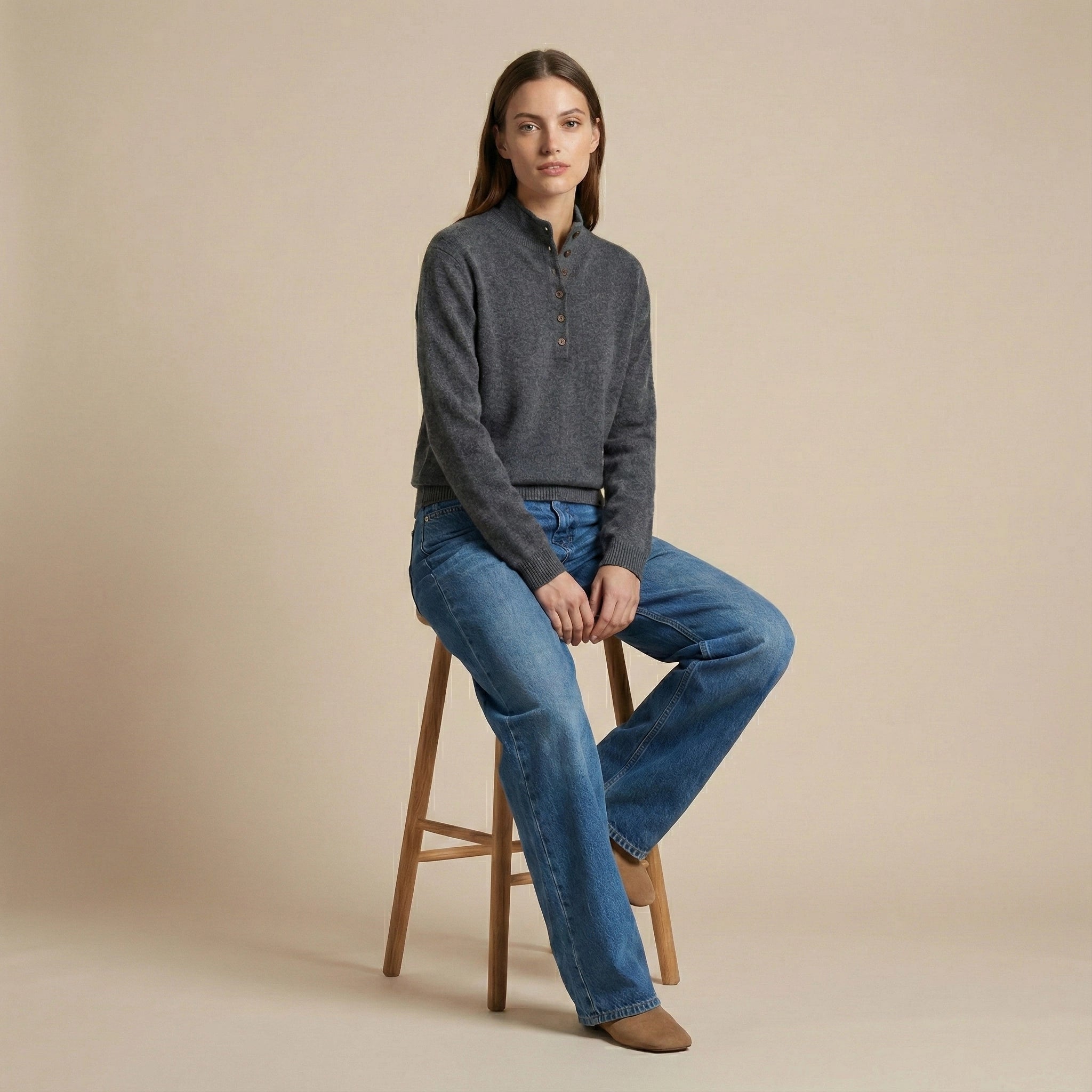 Woman sitting on a wooden stool wearing a gray sweater and blue jeans against a beige background