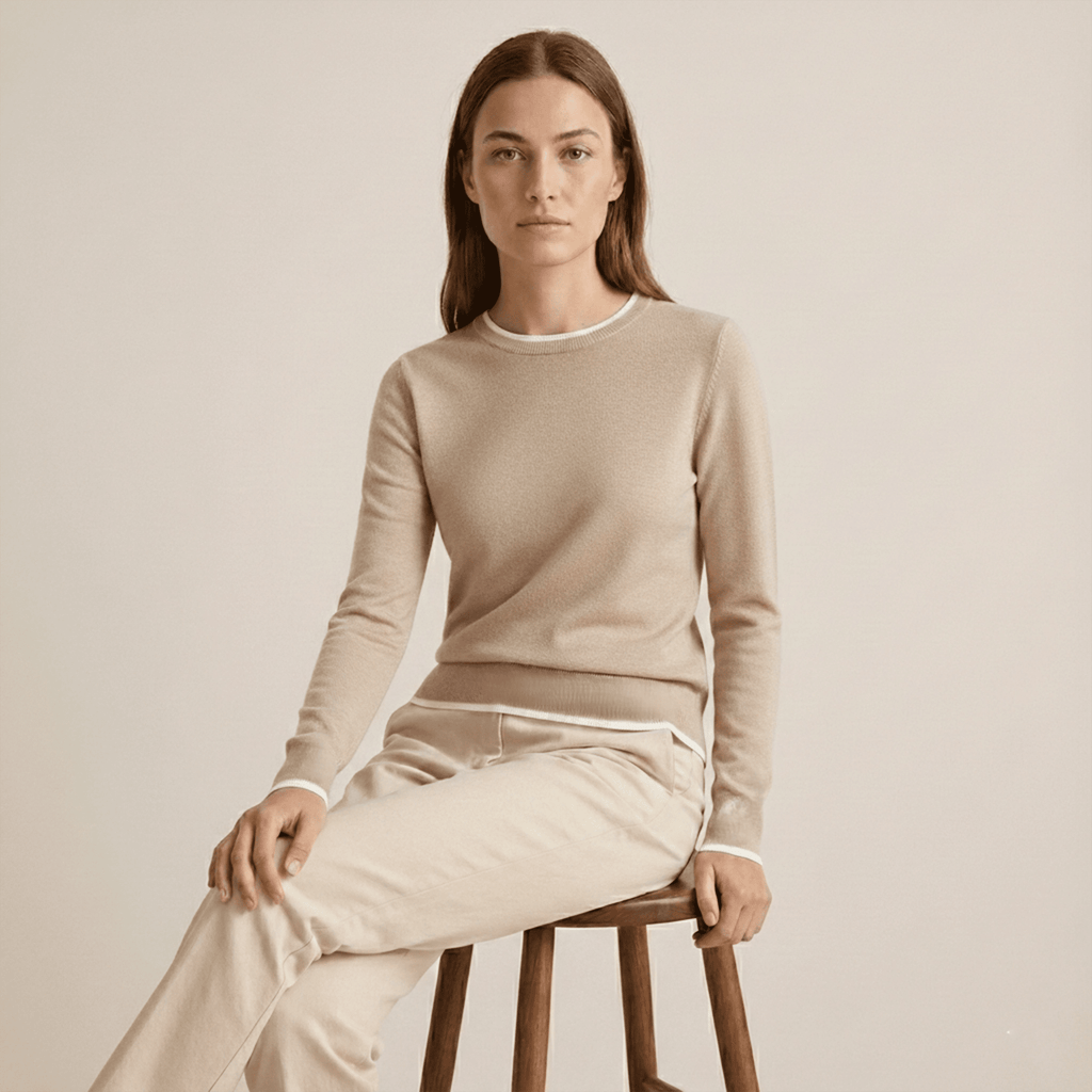 Woman wearing a beige sweater and pants sitting on a wooden stool against a plain background