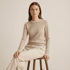 Woman wearing a beige sweater and pants sitting on a wooden stool against a plain background