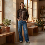 Woman in a brown sweater and blue jeans standing in a sunlit room with plants and furniture.