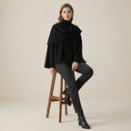 Woman wearing a black poncho sitting on a wooden stool against a beige background