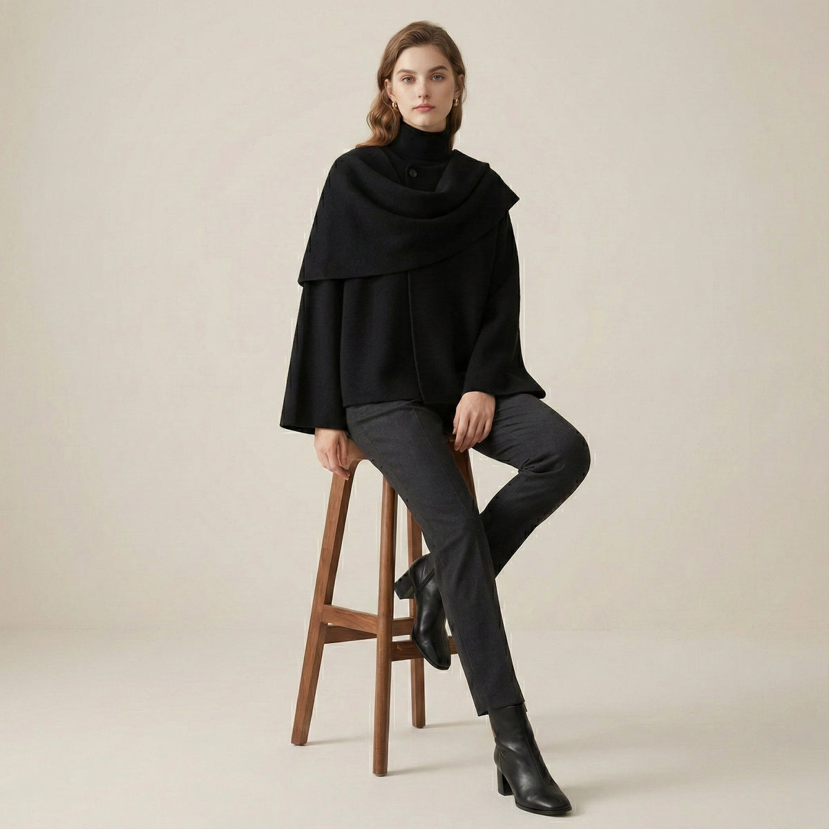 Woman wearing a black poncho sitting on a wooden stool against a beige background