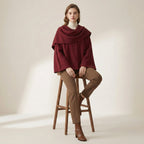 Woman wearing a burgundy poncho and brown pants sitting on a wooden stool against a plain background