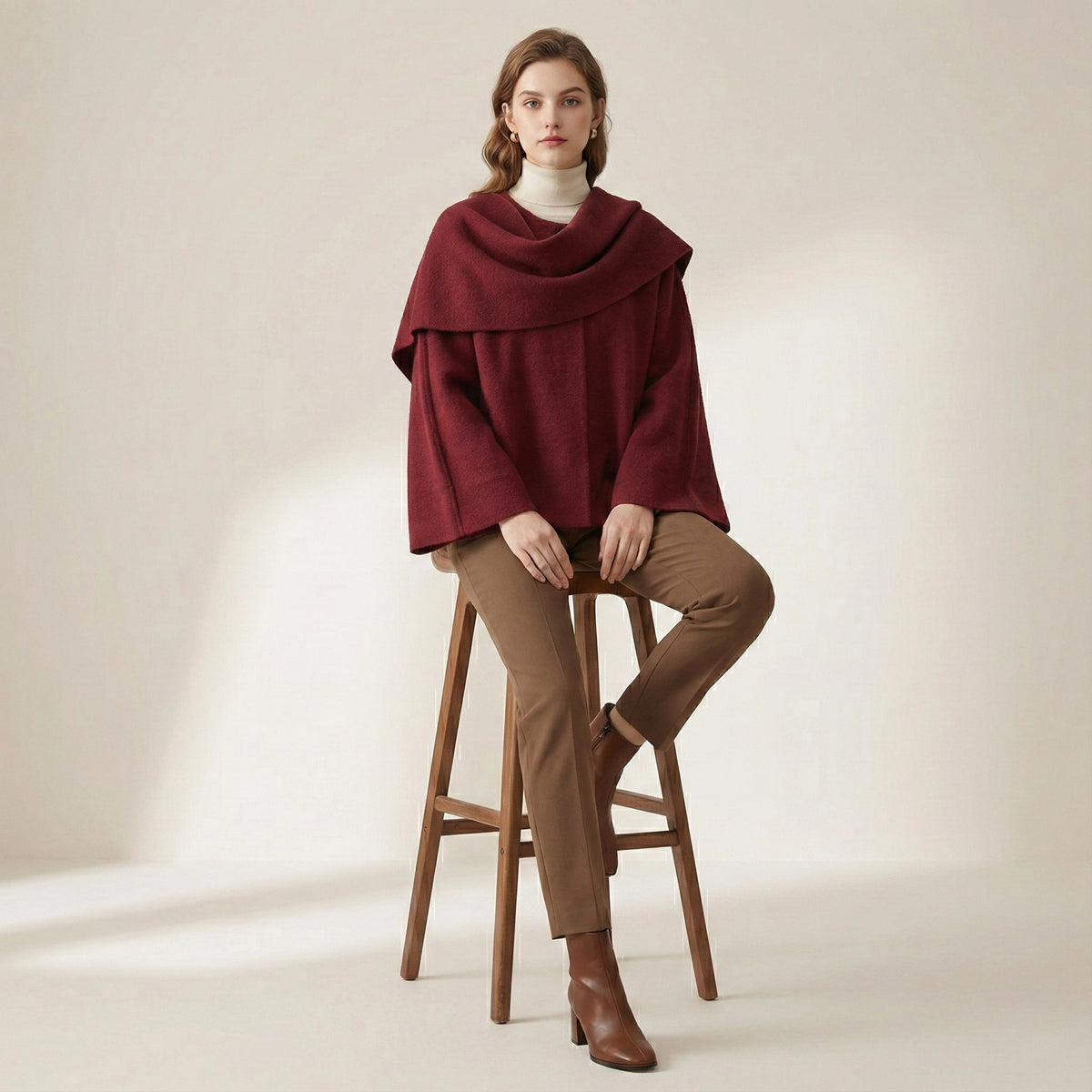 Woman wearing a burgundy poncho and brown pants sitting on a wooden stool against a plain background