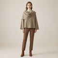 Woman wearing a beige cape over a white turtleneck and brown pants on a beige background
