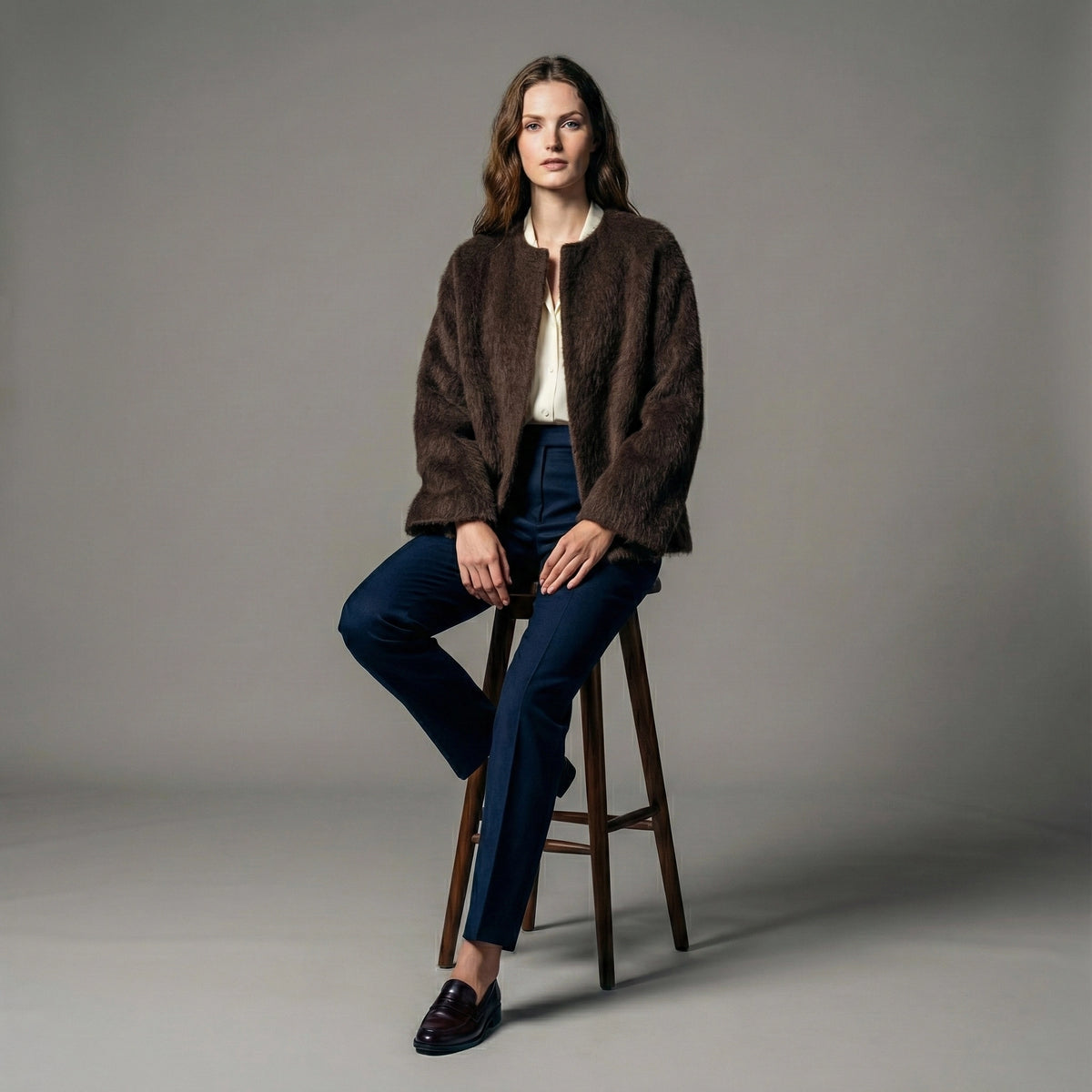 Woman sitting on a stool wearing a brown jacket and blue jeans against a gray background