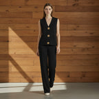 Woman wearing a black sleeveless blazer with gold buttons and matching pants on a gray background