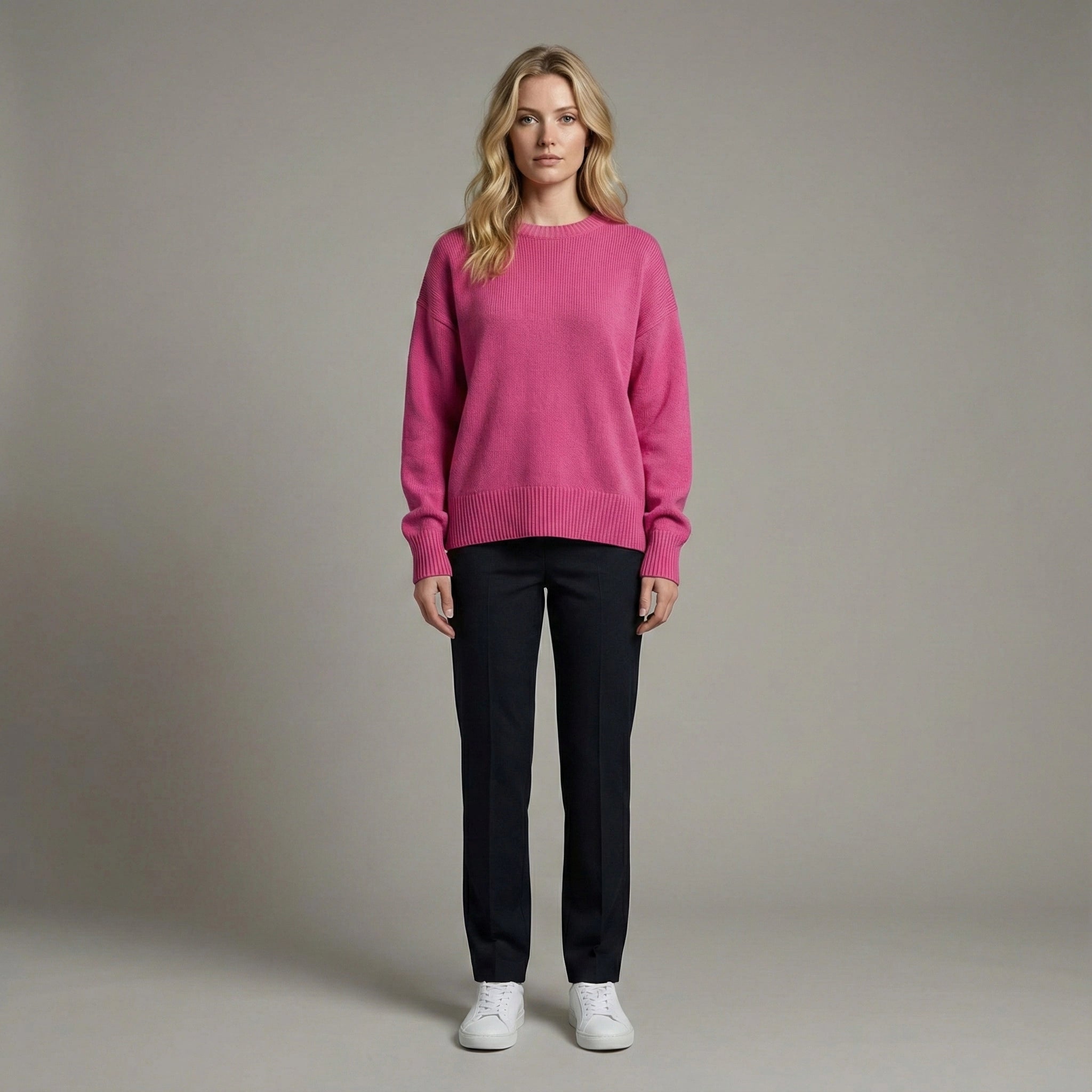 Woman wearing a pink sweater, black pants, and white shoes against a plain background
