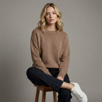 Woman wearing a brown sweater and black pants sitting on a wooden stool against a gray background