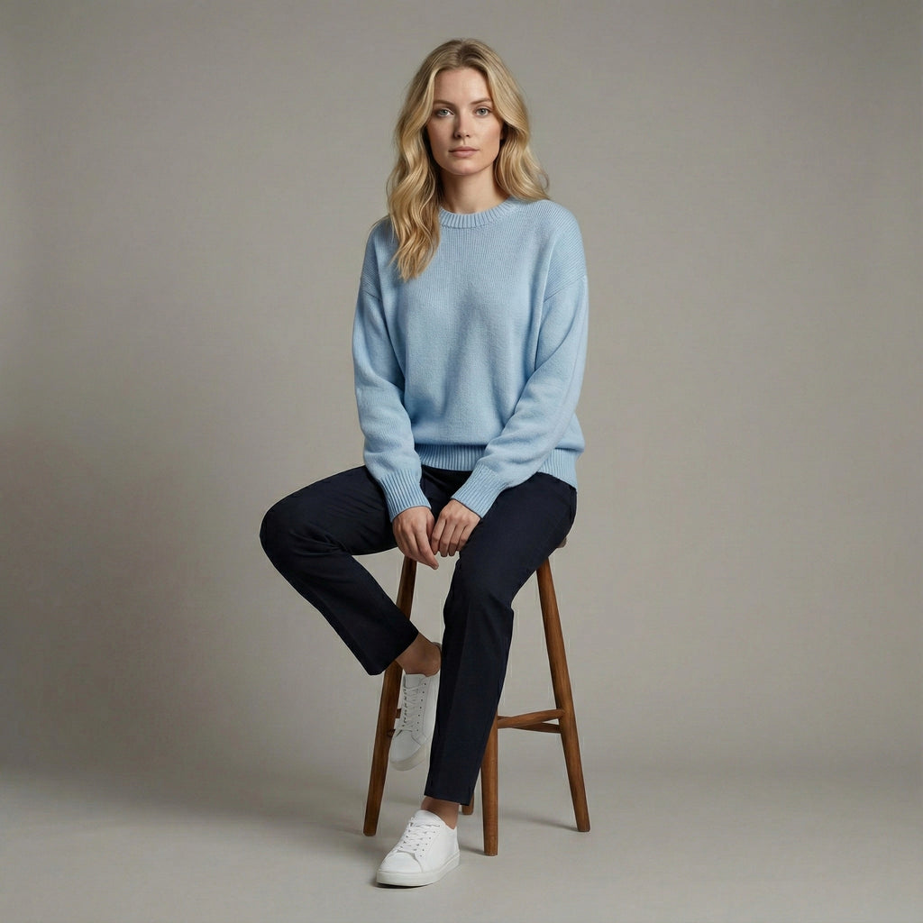 Woman sitting on a wooden stool wearing a light blue sweater and dark pants against a neutral background