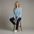 Woman sitting on a wooden stool wearing a light blue sweater and dark pants against a neutral background