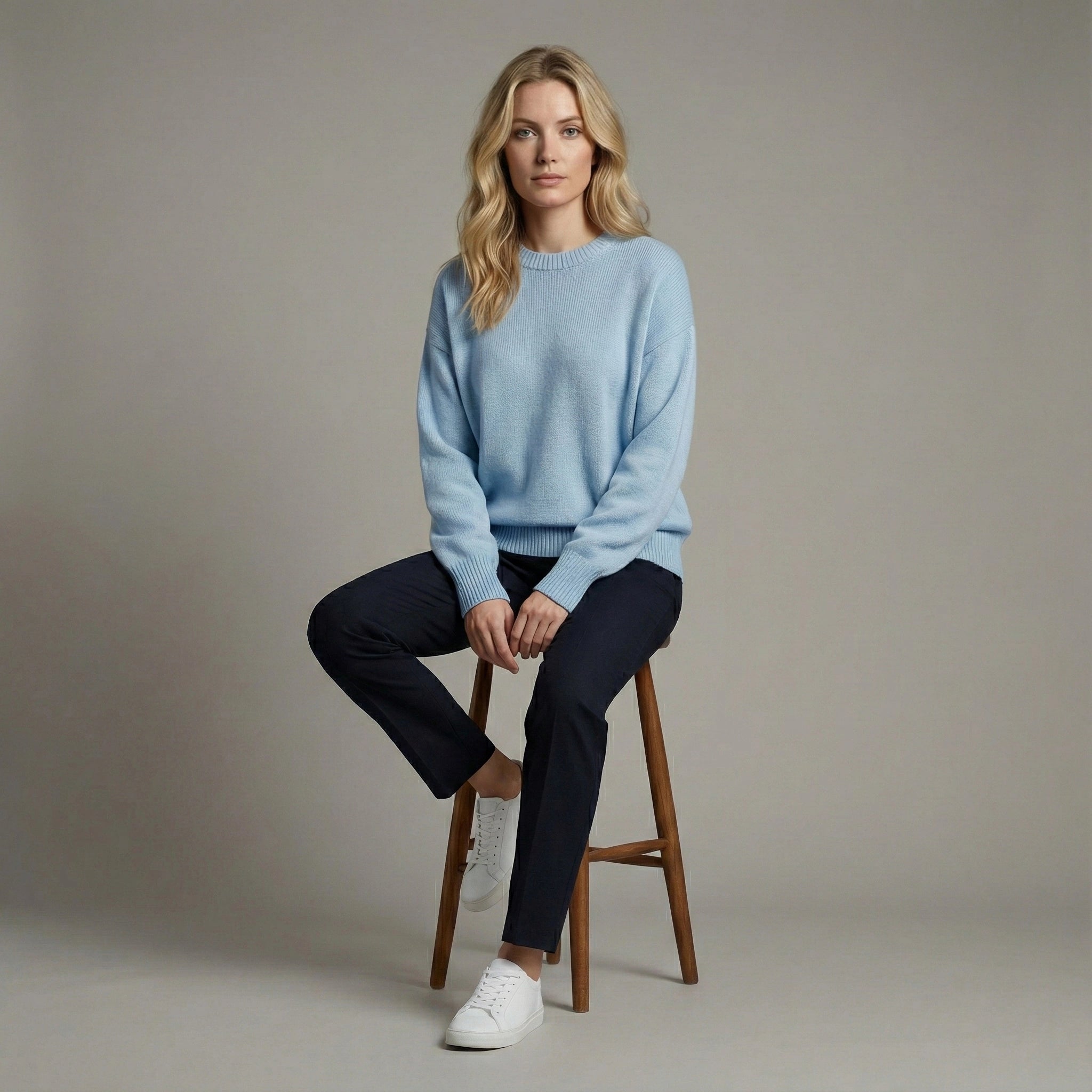 Woman sitting on a wooden stool wearing a light blue sweater and dark pants against a neutral background