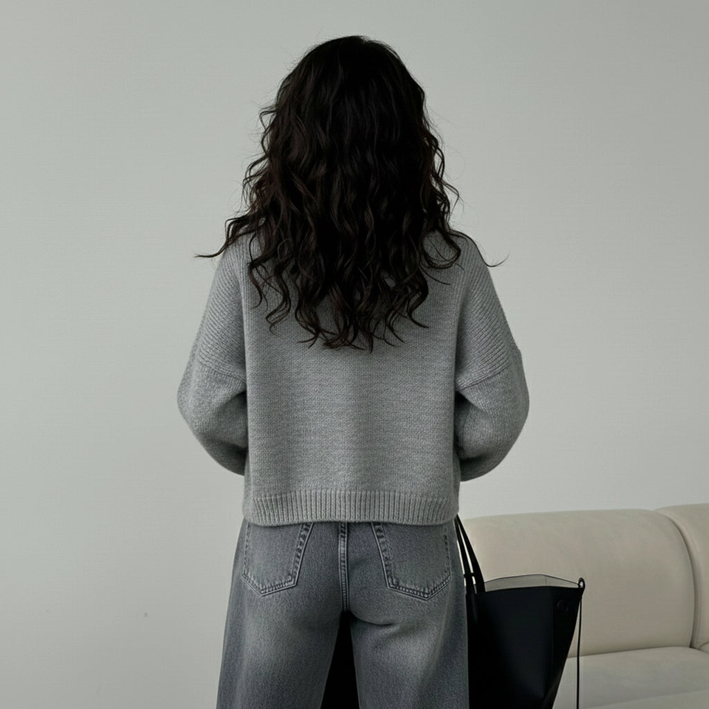 Person wearing a gray sweater and jeans standing in front of a beige sofa with a black handbag.