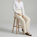 Person wearing a white shirt and beige pants sitting on a wooden stool against a plain background