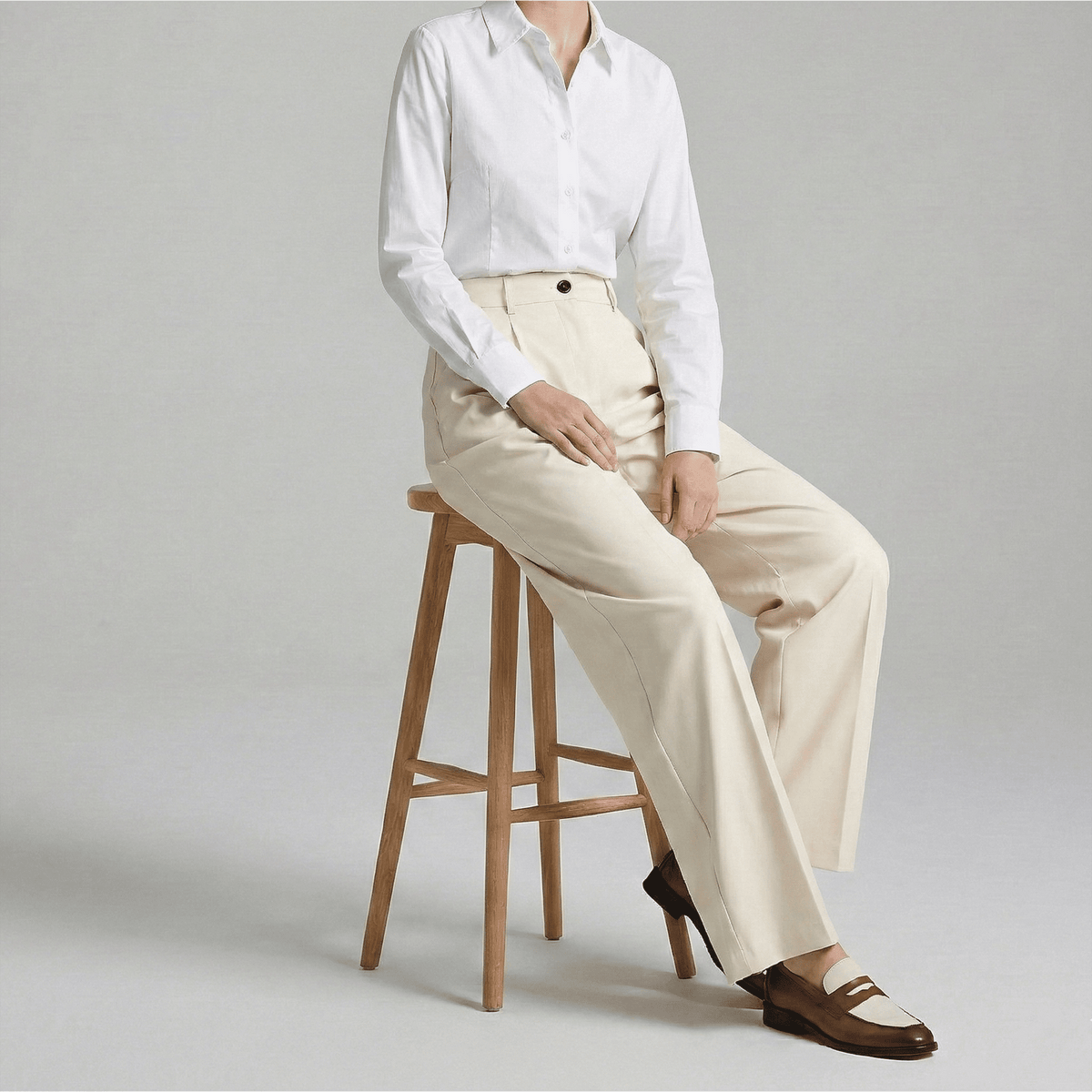 Person wearing a white shirt and beige pants sitting on a wooden stool against a plain background