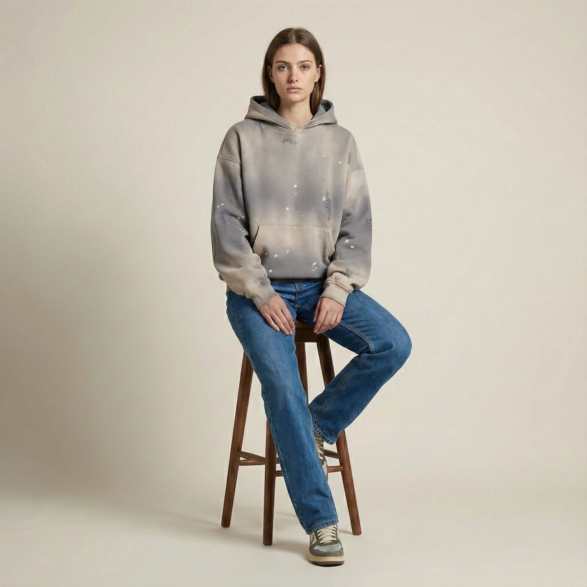 Person wearing a gray tie-dye hoodie and blue jeans sitting on a wooden stool against a beige background
