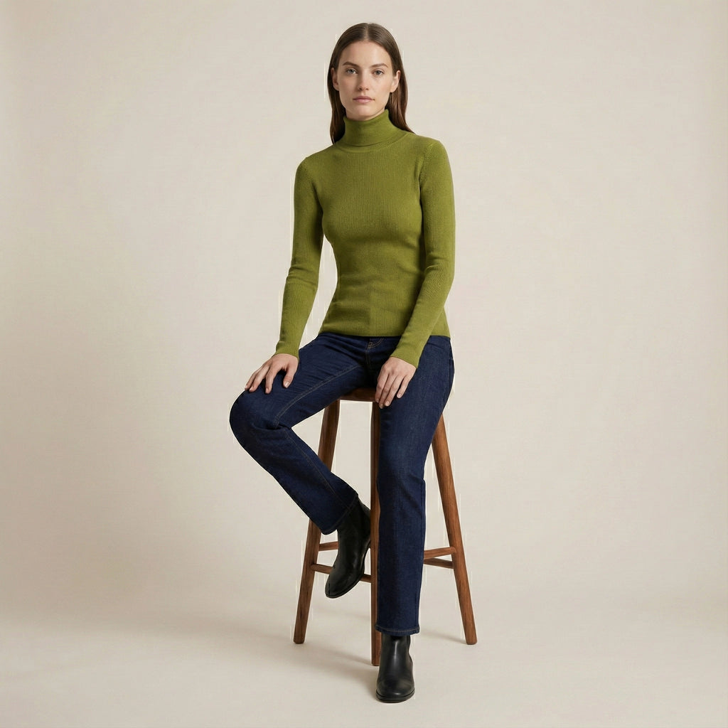 Woman wearing a green turtleneck sweater and blue jeans sitting on a wooden stool against a beige background
