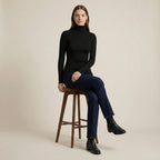 Woman sitting on a wooden stool wearing a black turtleneck and dark jeans against a beige background