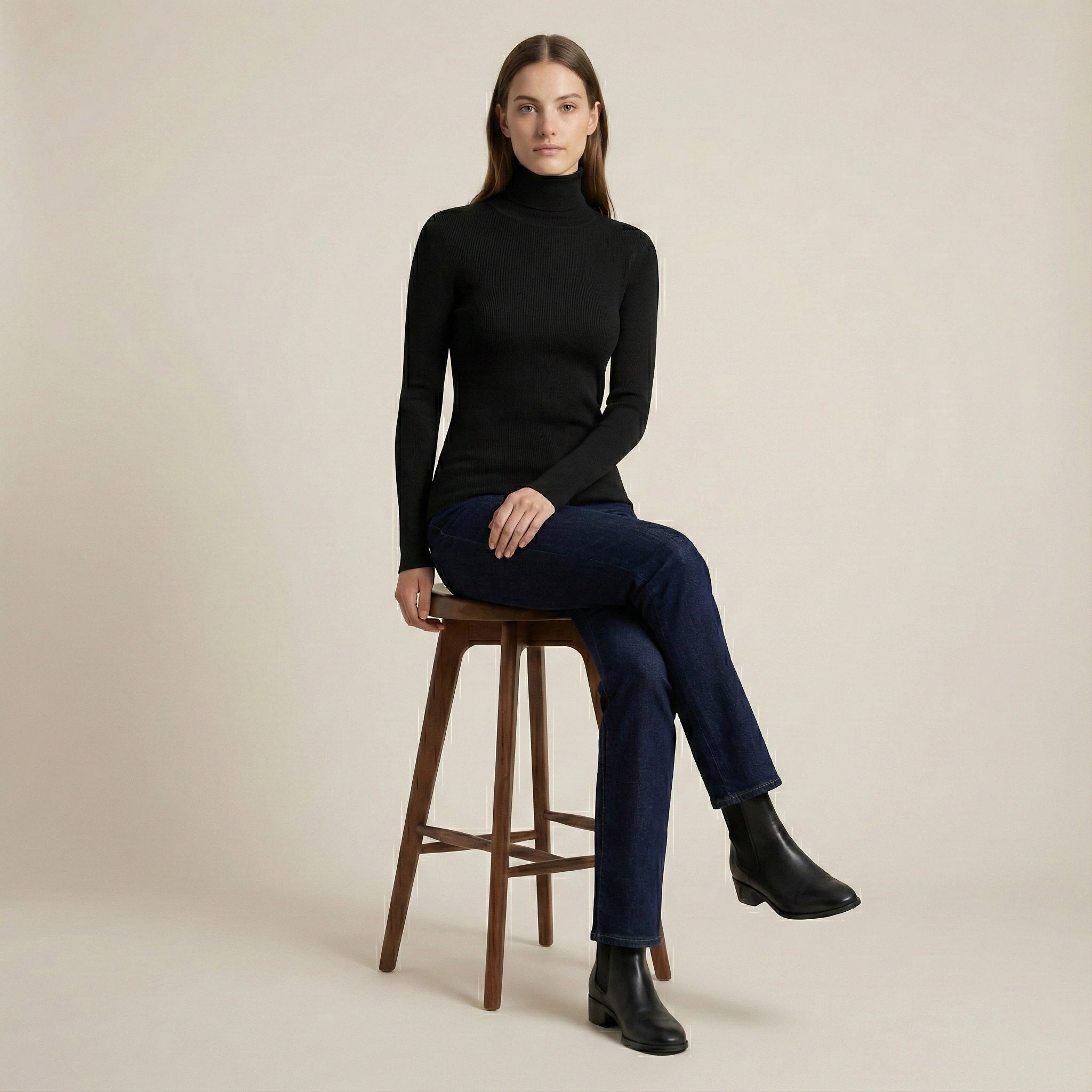 Woman sitting on a wooden stool wearing a black turtleneck and dark jeans against a beige background