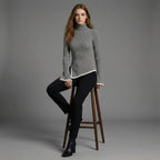 Woman wearing a gray turtleneck sweater and black pants sitting on a wooden stool against a gray background