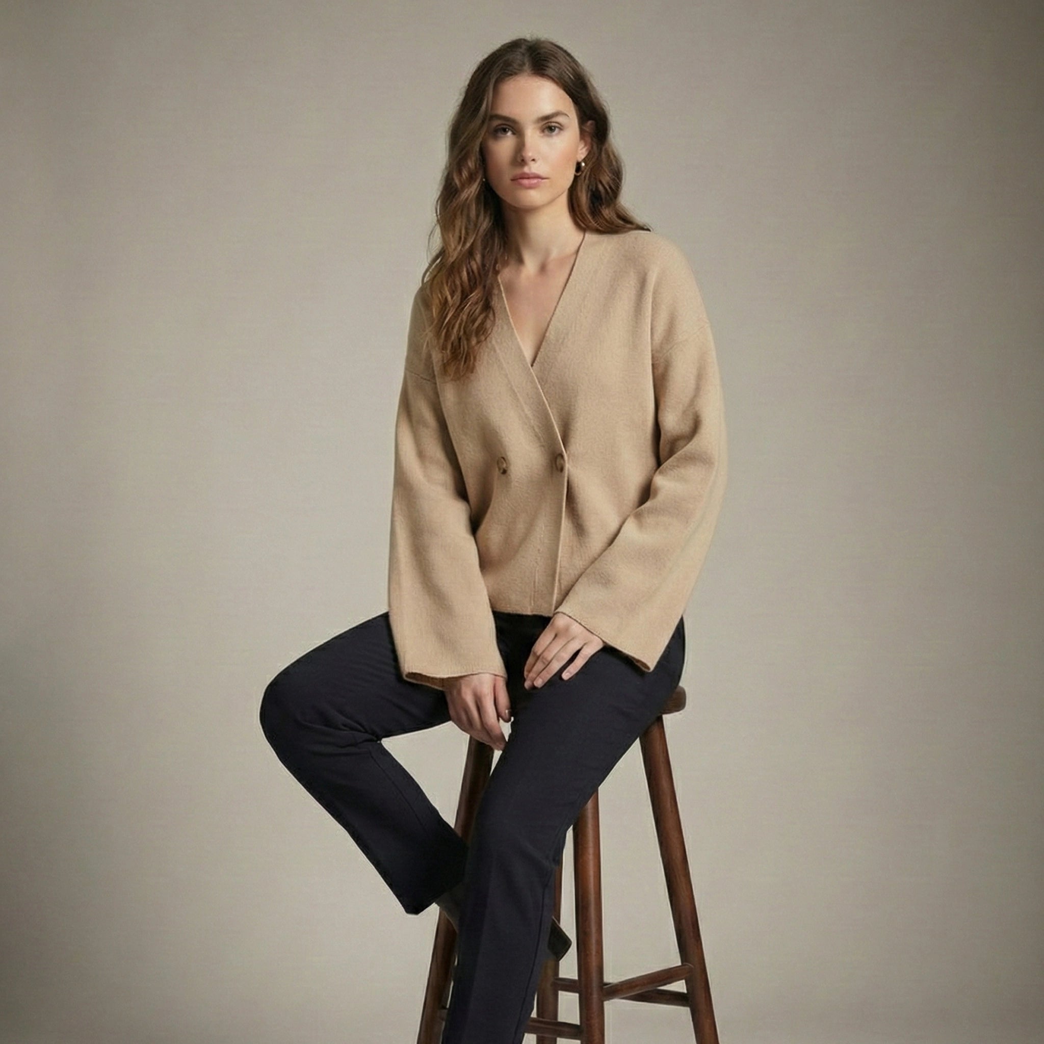 Woman wearing a beige cardigan and black pants sitting on a wooden stool against a plain background