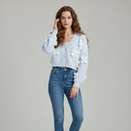 Woman wearing a light blue cardigan with white floral embellishments and blue jeans on a gray background