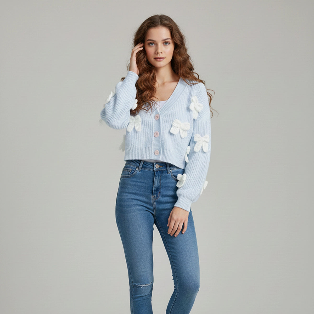 Woman wearing a light blue cardigan with white floral embellishments and blue jeans on a gray background