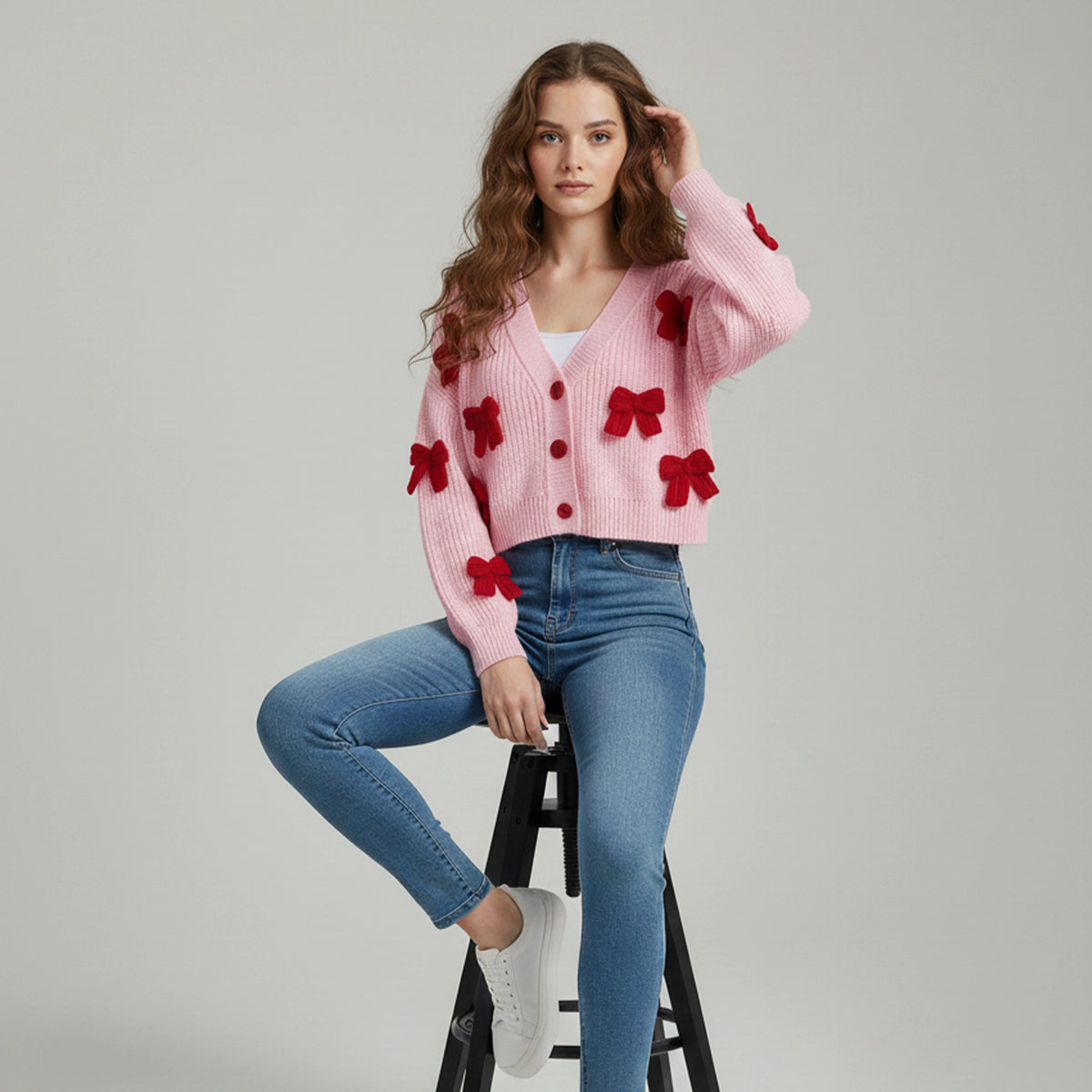 Woman wearing a pink cardigan with red bow details sitting on a black stool against a gray background