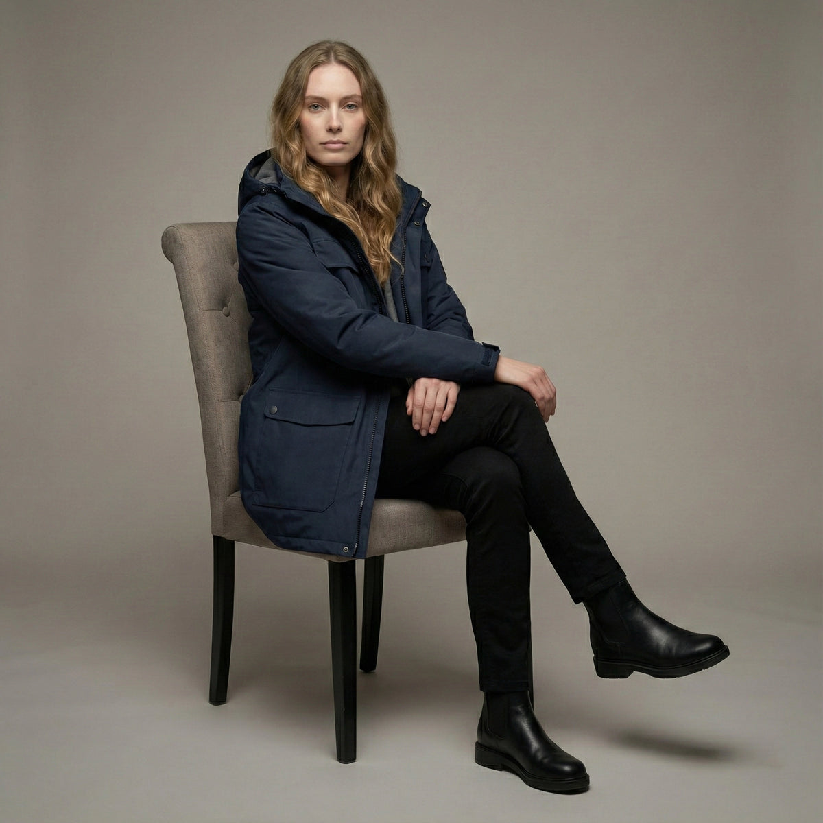 Woman sitting on a chair wearing a navy coat and black pants against a plain background