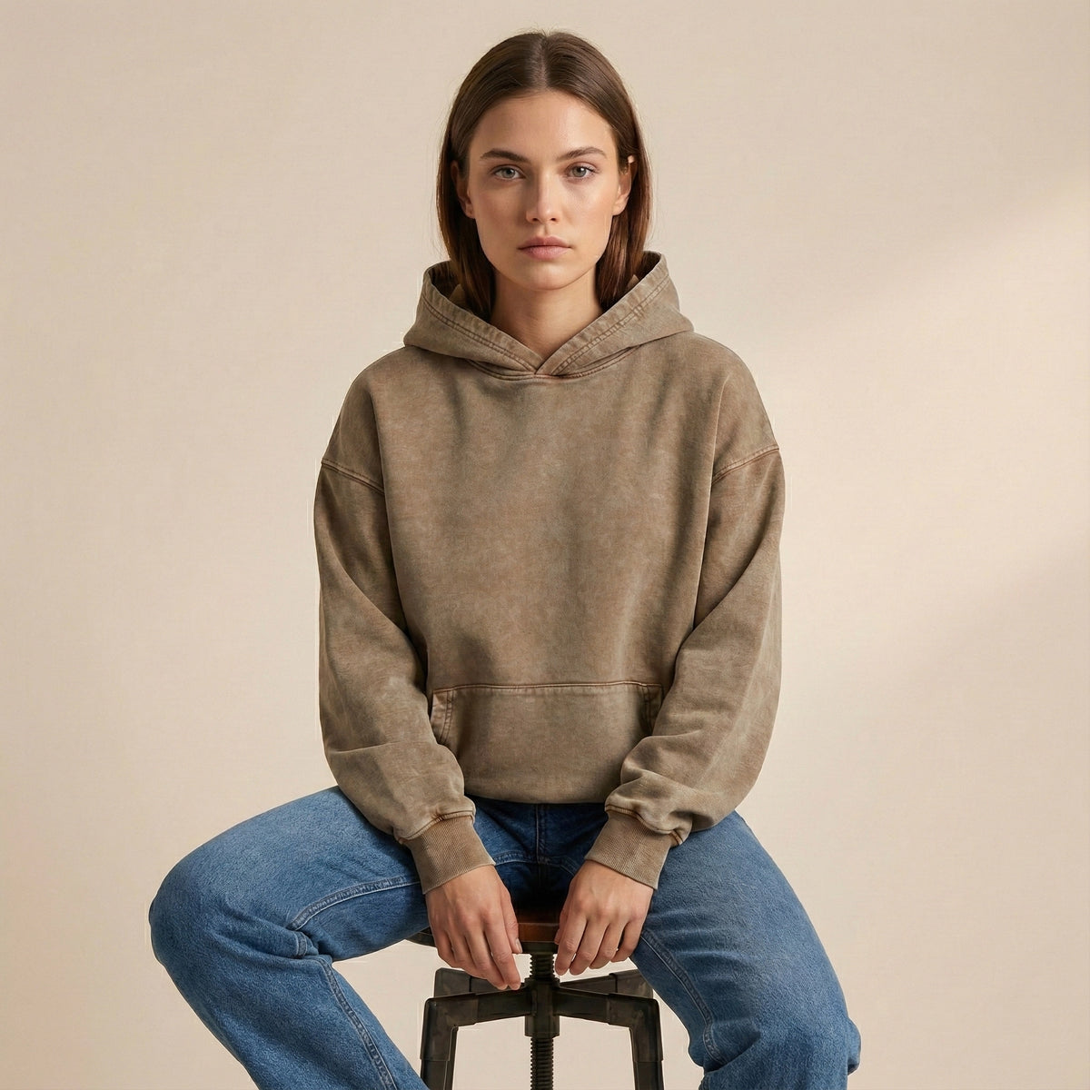 Person wearing a brown hoodie and blue jeans sitting on a stool against a beige background