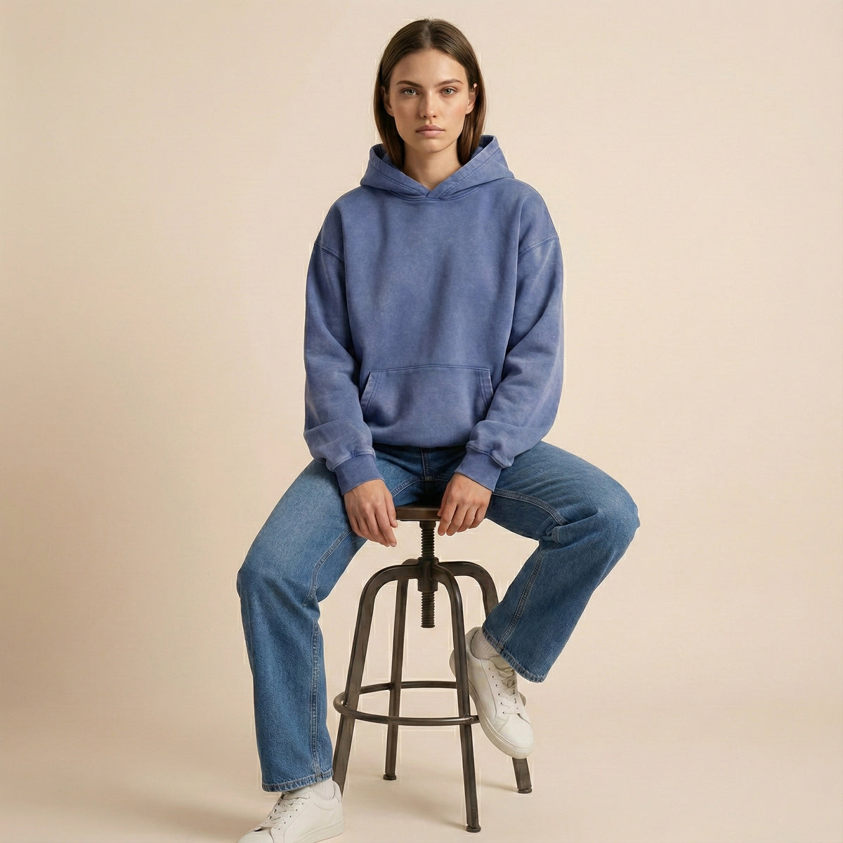 Person wearing a blue hoodie and jeans sitting on a stool against a beige background