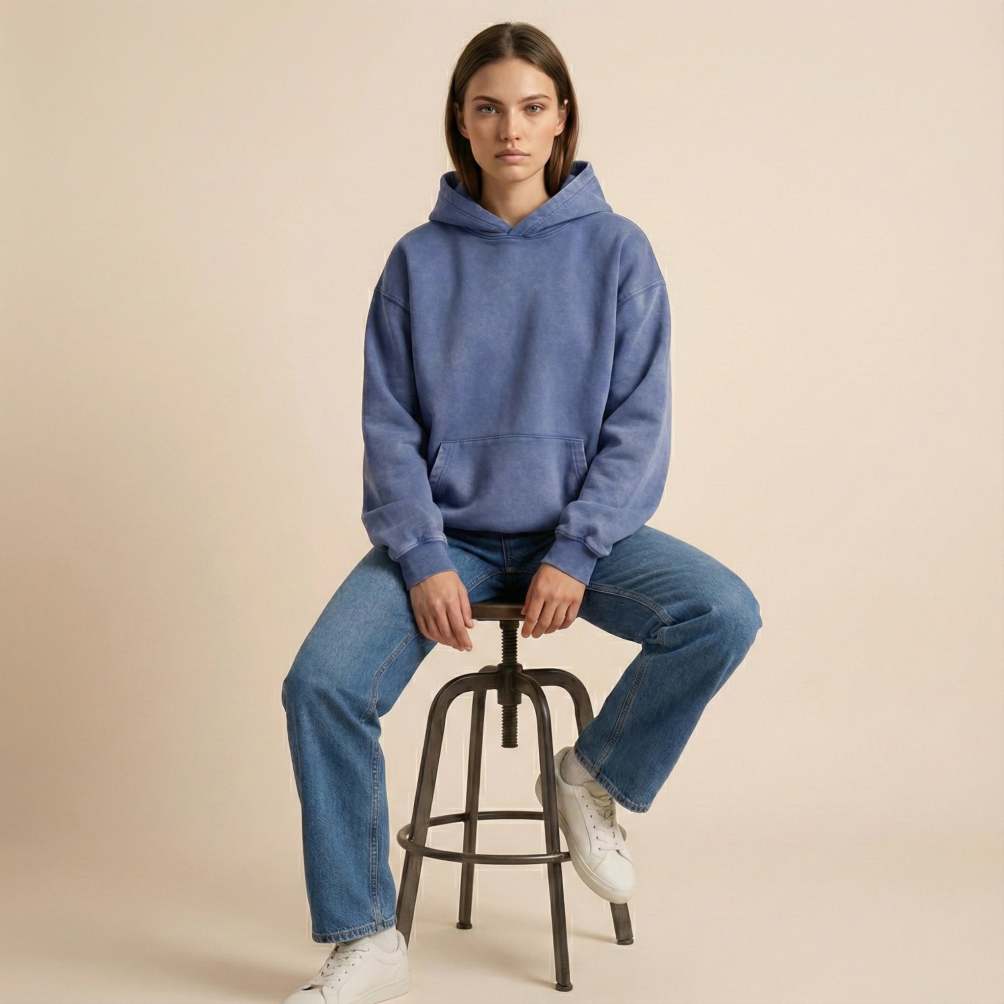 Person wearing a blue hoodie and jeans sitting on a stool against a beige background