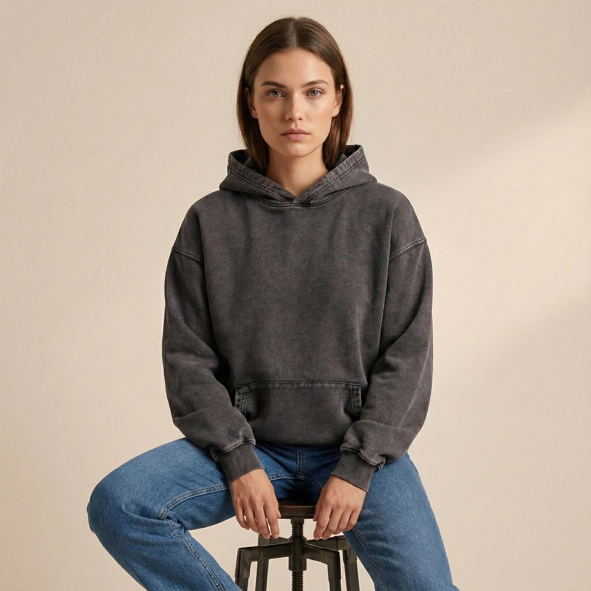 Person wearing a gray hoodie and blue jeans sitting on a stool against a beige background