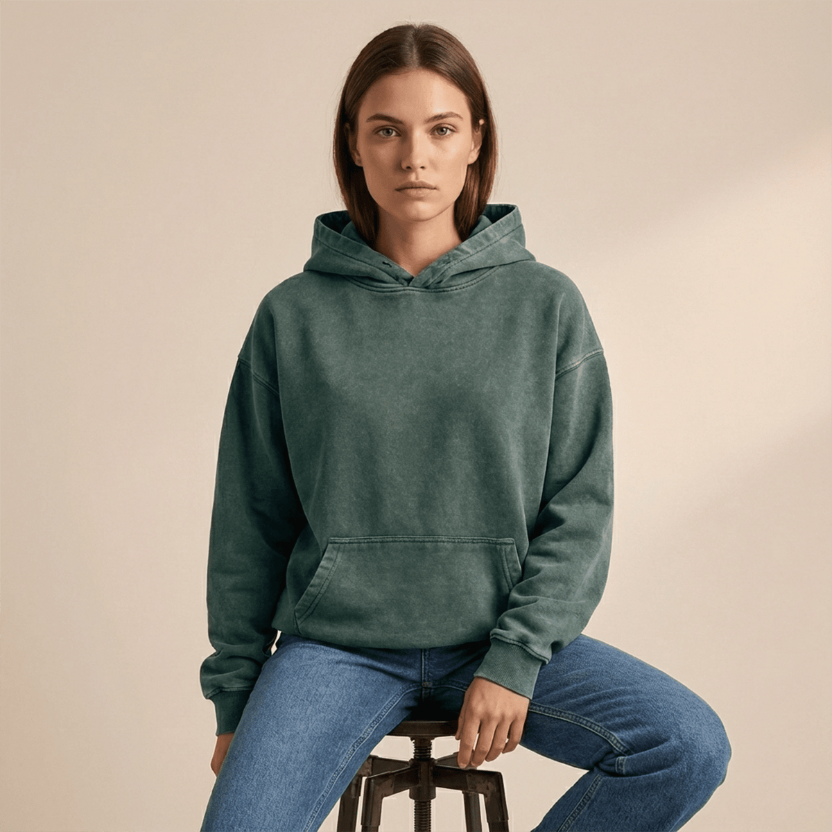 Person wearing a green hoodie and blue jeans sitting on a stool against a beige background