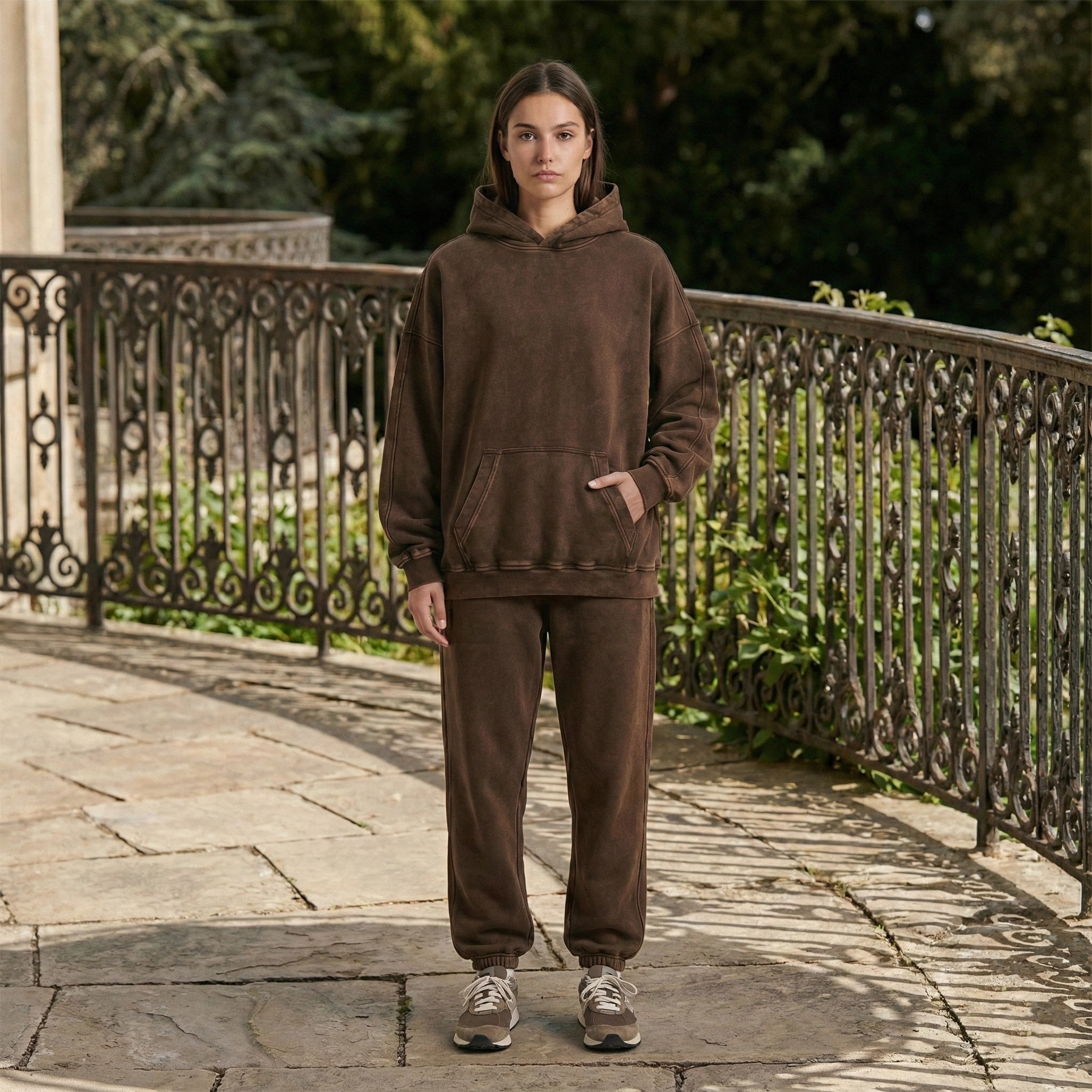 Person wearing a brown hoodie and pants standing on a stone pathway with greenery in the background